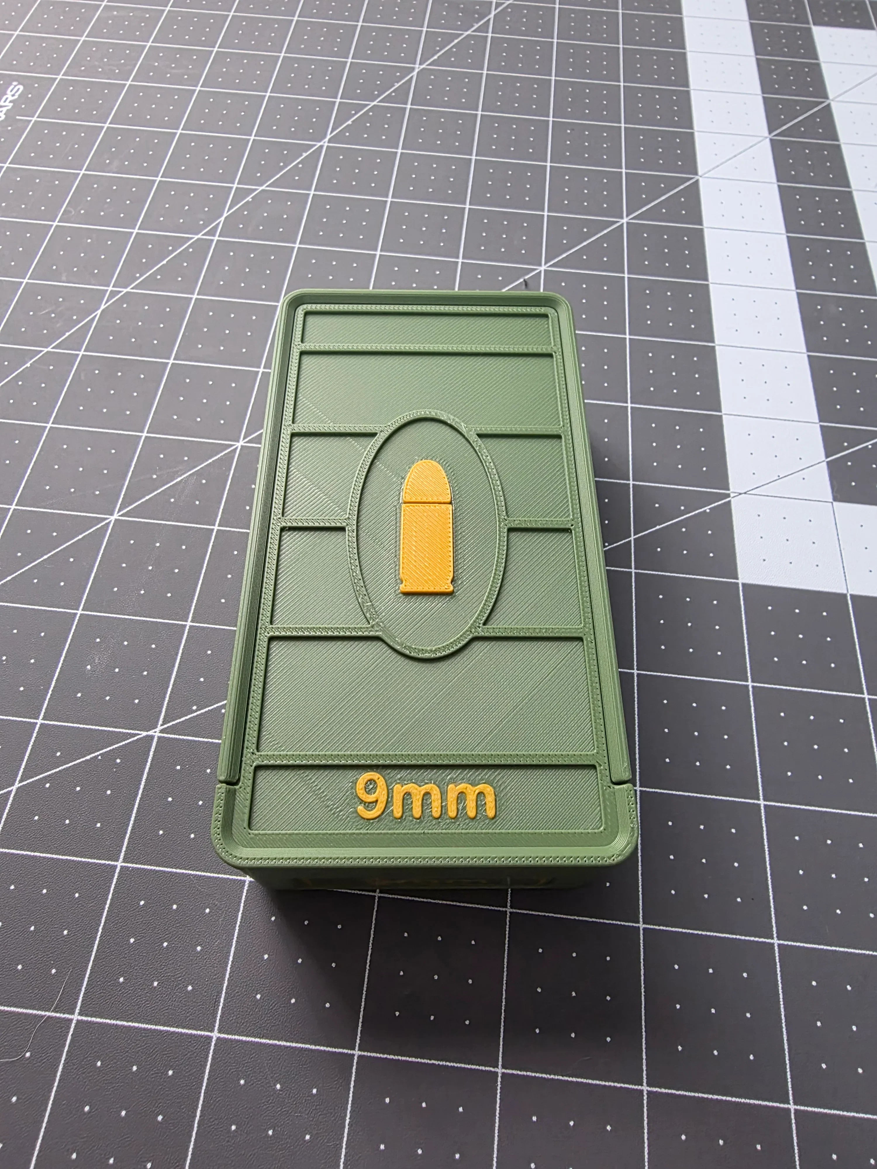 Green 3D-printed 9mm ammo storage box with yellow bullet icon on a cutting mat