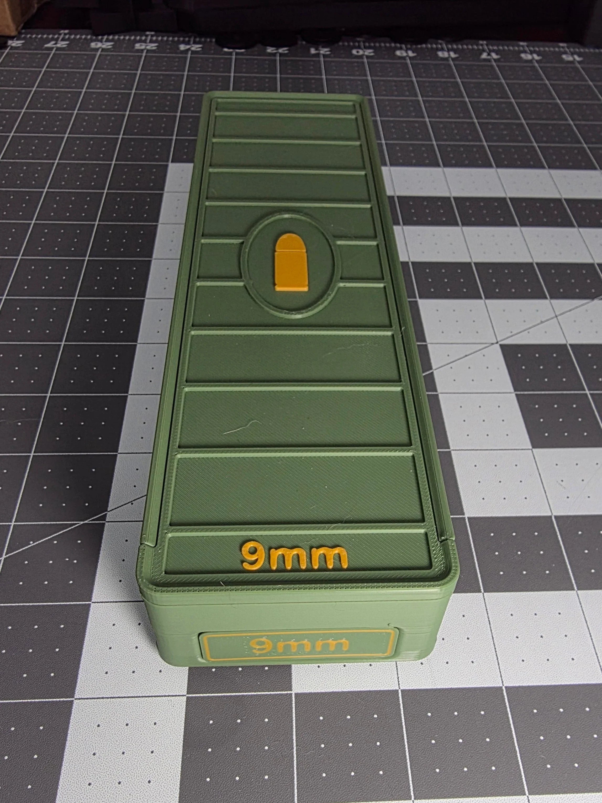 Green 3D-printed 9mm ammo storage box with yellow lettering on a grid mat.