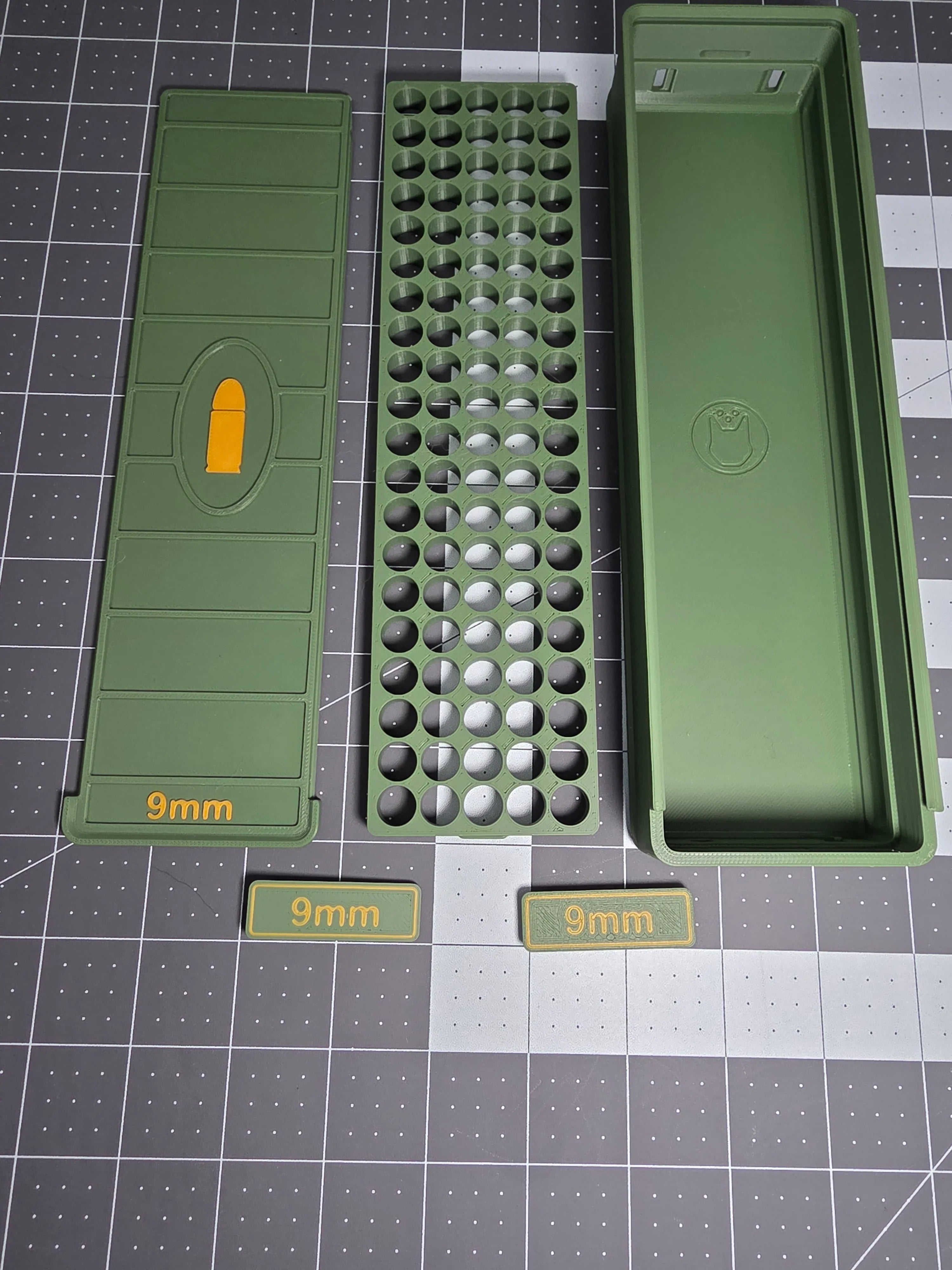 Green 3D-printed 9mm ammo storage box disassembled, showing tray and labeled parts on grid mat.