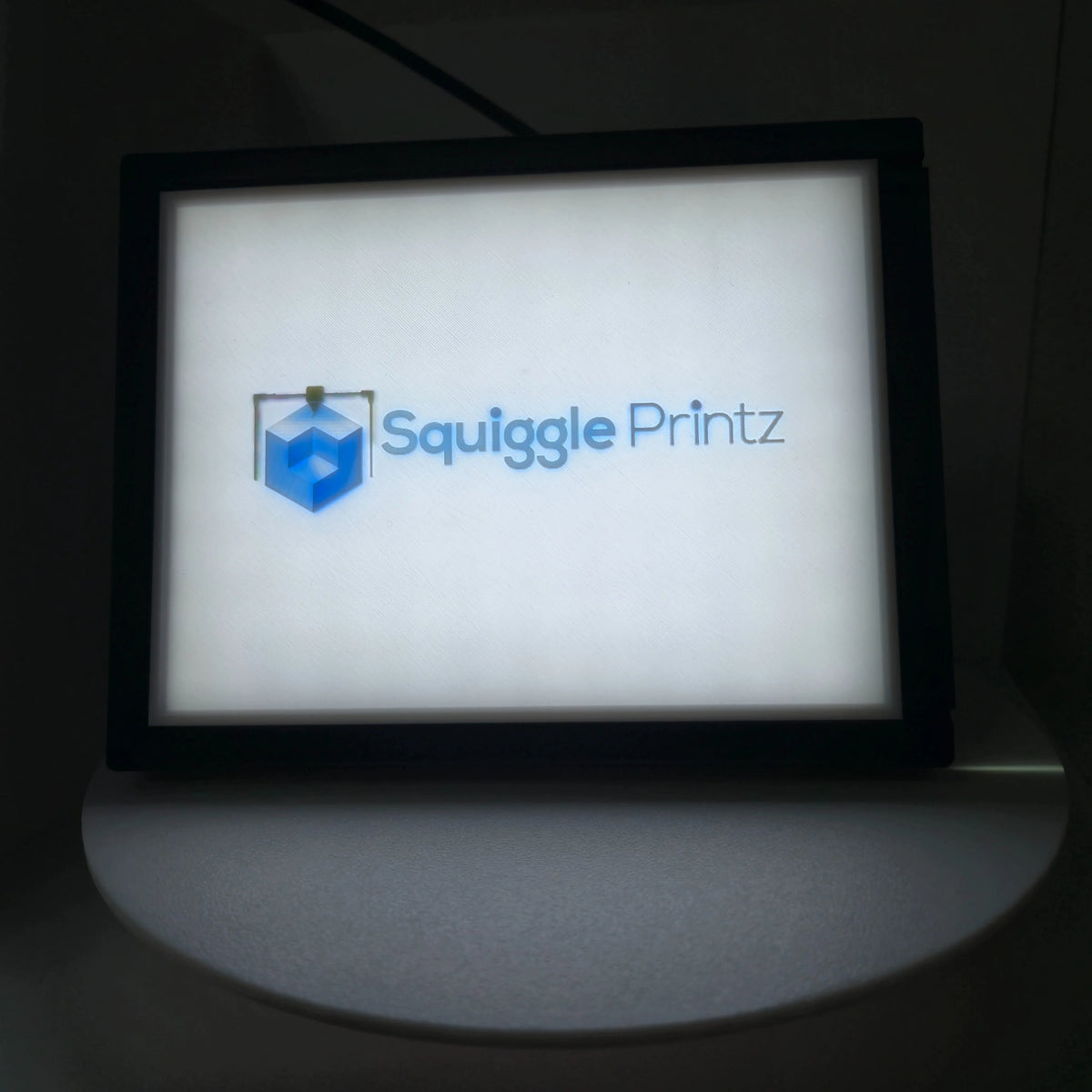 Lithophane lit from behind in its display frame, showing a full-color SquigglePrintz logo