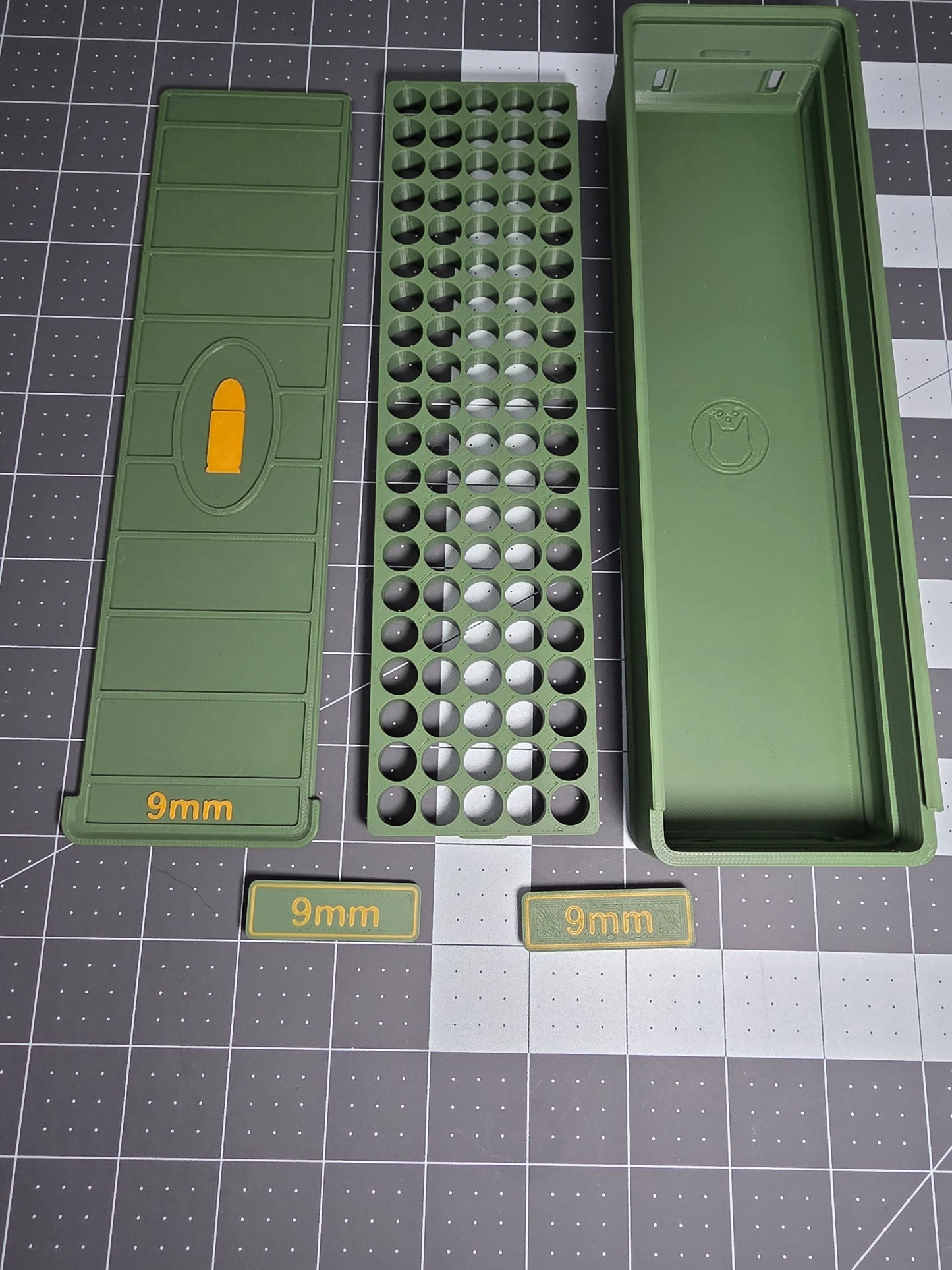 Disassembled green 3D-printed 9mm 100-round ammo storage box showing tray and lid on grid mat.